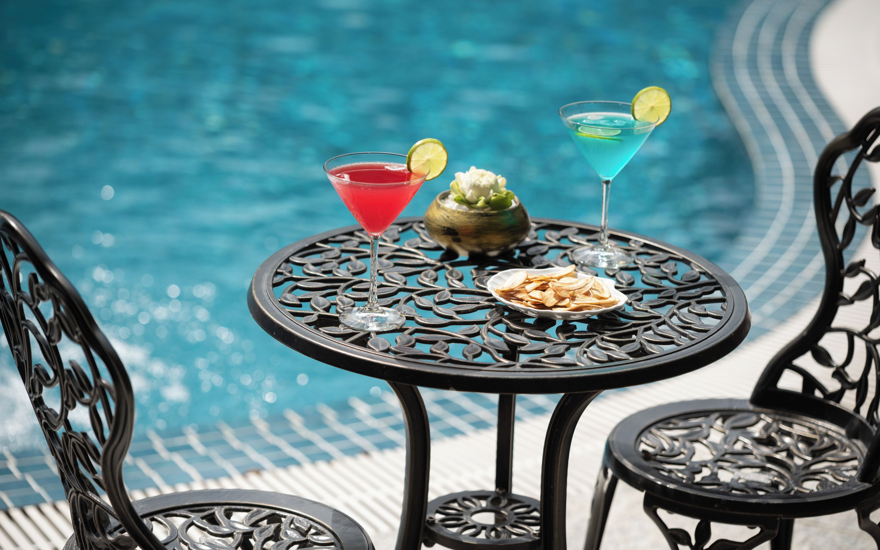 A poolside table with two drinks and a bowl of snacks, surrounded by chairs.