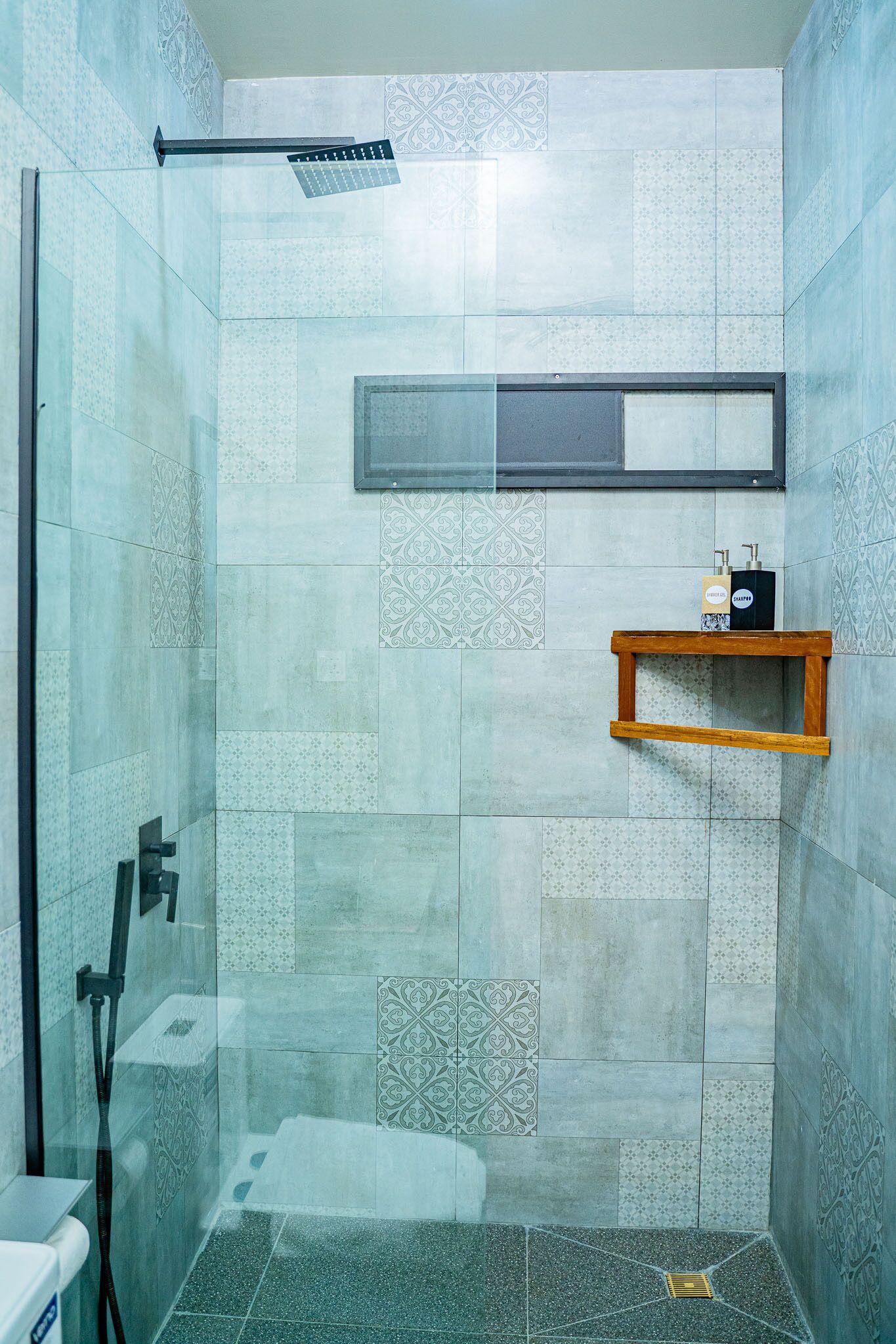 Un área de ducha con puerta de vidrio, ducha, repisa con artículos de aseo y azulejos estampados.