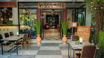 A restaurant entrance with a 'WELCOME' sign, outdoor seating with tables and chairs, and a 'HAPPY 5 PM ZEN Hour' sign.
