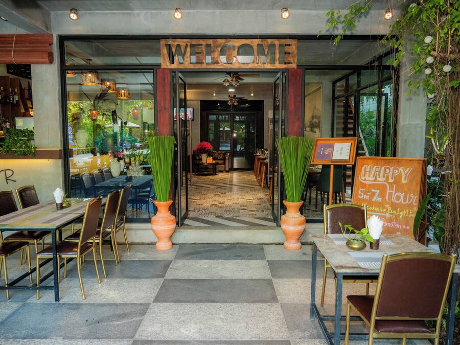 Een restaurantingang met een 'WELCOME' bord, terras met tafels en stoelen, en een 'HAPPY 5 PM ZEN Hour' bord.