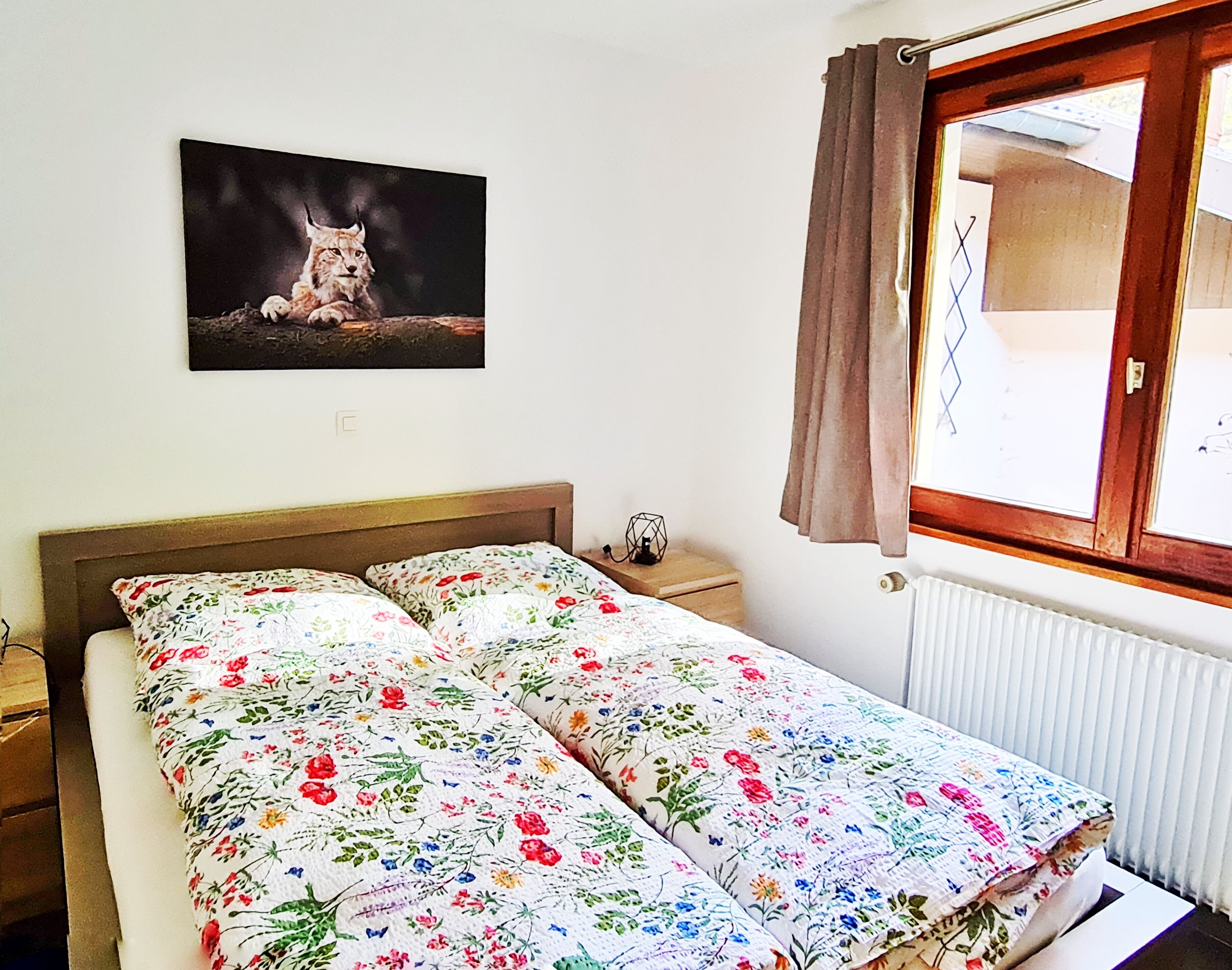 Ein Schlafzimmer mit floralem Bettbezug, einem Nachttisch mit Lampe, einem gerahmten Bild an der Wand und einem Fenster mit Vorhängen.