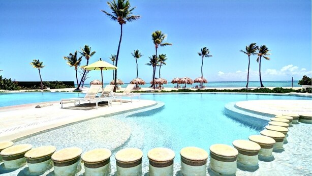 A pool with clear blue water, surrounded by lounge chairs and umbrellas, with palm trees and a beach in the background.