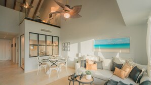 A spacious living area with a high ceiling, a dining area with white furniture, and a large window with a painting of a beach.