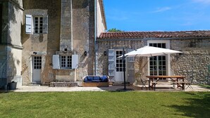 A stone building with a tiled roof, a covered patio with a table and chairs, and a grassy yard. - Beautiful house with swimming pool on the banks of the Charente river (Bourg-Charente)