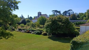 A well-maintained grassy area with a variety of trees and shrubs, a clear sky, and a building with a bell tower in the background. - Beautiful house with swimming pool on the banks of the Charente river (Bourg-Charente)