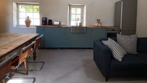 A compact kitchen with a wooden table, two windows, a built-in oven, and a dark sofa with a patterned pillow. - Beautiful house with swimming pool on the banks of the Charente river (Bourg-Charente)