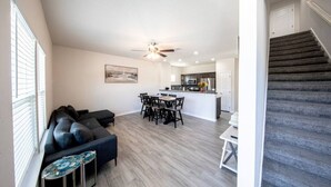 A modern living room with a dark-colored sofa, a dining table with chairs, and a kitchen area with a white countertop and dark cabinets.
