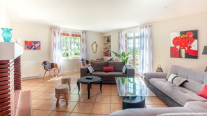 Un salón amplio con un sofá gris, una mesita de centro y un sillón rojo. Hay un ventanal con cortinas, una planta y obras de arte en las paredes.