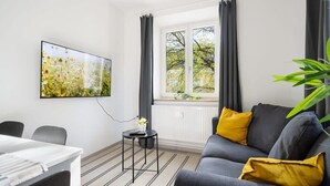 A modern living room with a grey sofa, a small side table with a plant, a dining table, and a large window with curtains. - Apartment an der Theresienwiese (München)