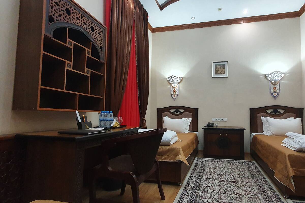 A hotel room with two single beds, a desk with a chair, a small table, a decorative wall piece, and a patterned rug.