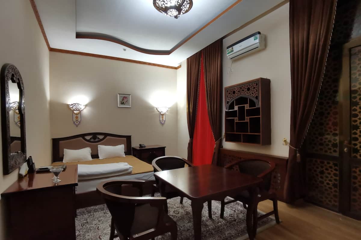 A hotel room with a bed, bedside table, chairs, a desk, a mirror, wall sconces, an air conditioning unit, and a patterned rug.