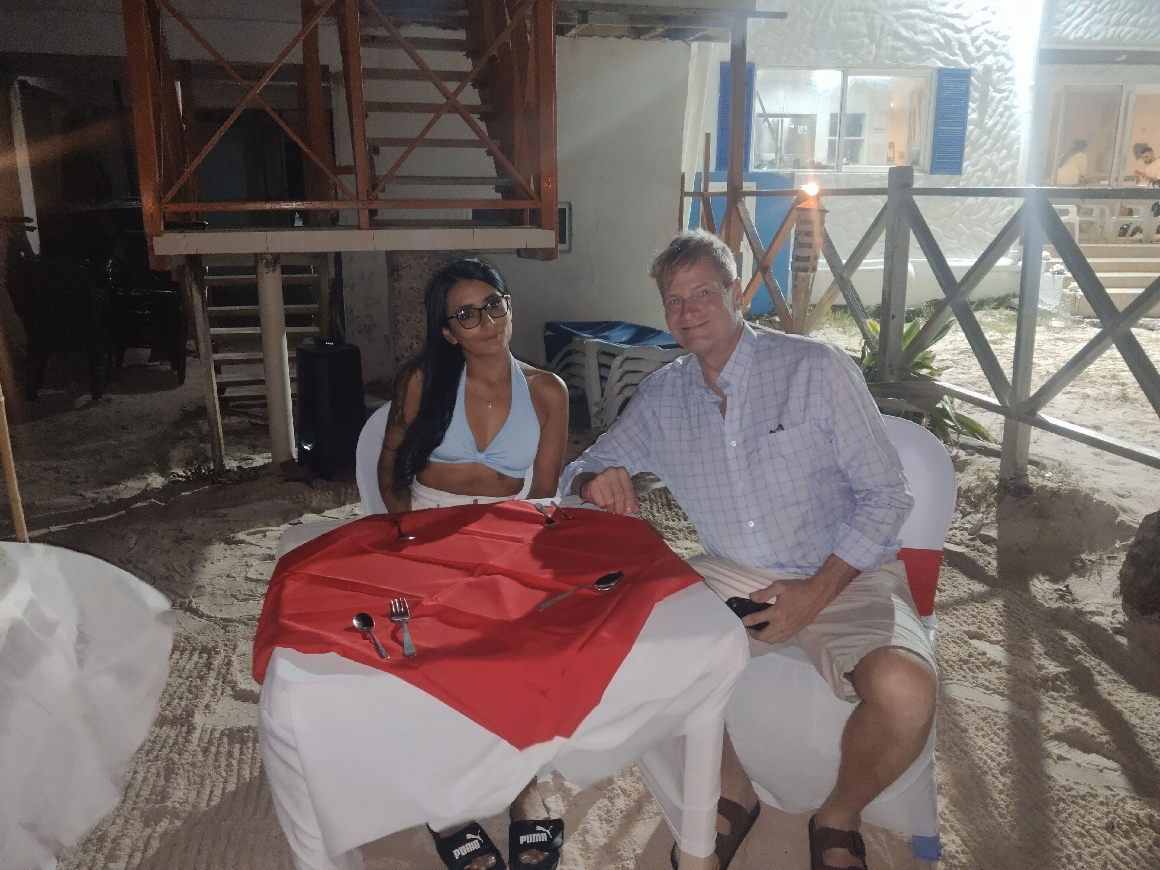 Dos personas sentadas en una mesa con mantel rojo y blanco, una estructura de madera al fondo y un área exterior de arena.