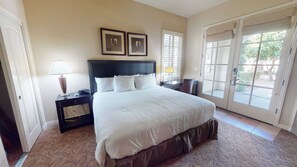 Quarto de hotel com uma cama grande, duas mesas de cabeceira, uma escrivaninha e uma cadeira. Há duas janelas com persianas, uma porta e vista para o exterior.