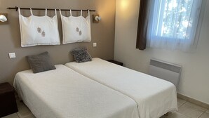 A bedroom with two beds, a nightstand, a wall-mounted shelf with towels, and a window with curtains.