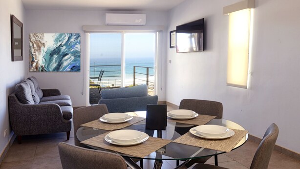 A modern living room with a glass table set for four, a grey sofa, and a large abstract painting. - Popotla Villa 203 (PLAYAS DE ROSARITO)