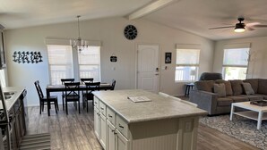 Une cuisine moderne dotée d’un îlot central, un coin repas avec une table et des chaises, un salon avec un canapé et une table basse, et un ventilateur de plafond.