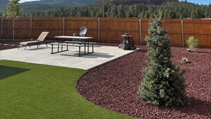 Un jardin avec une clôture en bois, un barbecue, une table de pique-nique et un petit arbre.