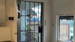 Ein Zimmer mit einer Schiebetür zum Balkon, ein Sofa mit einem strukturierten Zierkissen, ein kleiner Tisch und ein Fenster mit Blick auf einen Holenzaun und parkende Autos.