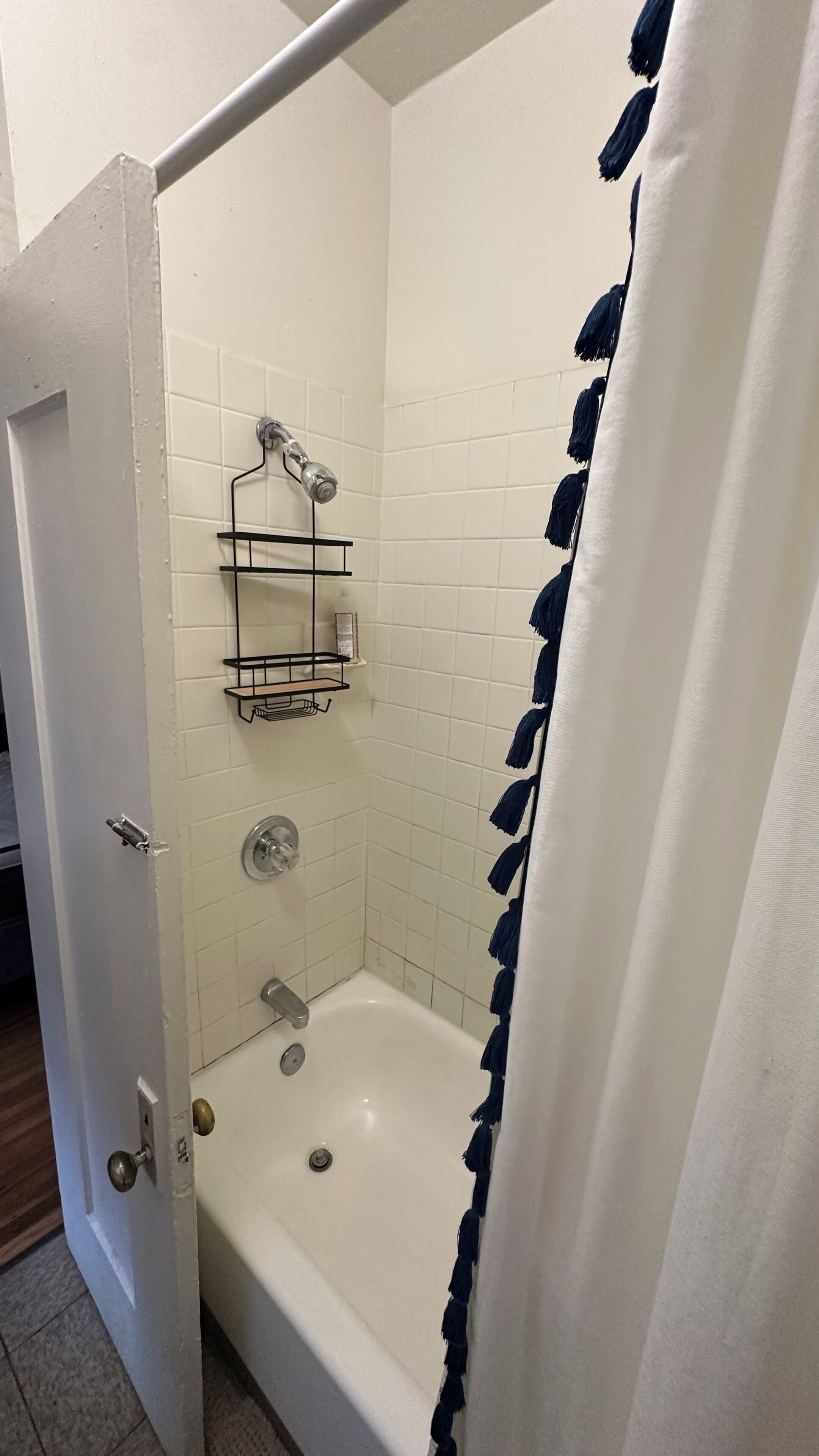 Ein kleines Badezimmer mit weißen Fliesen, einer Duschvorhang, einem Metallregal und einer Badewanne.