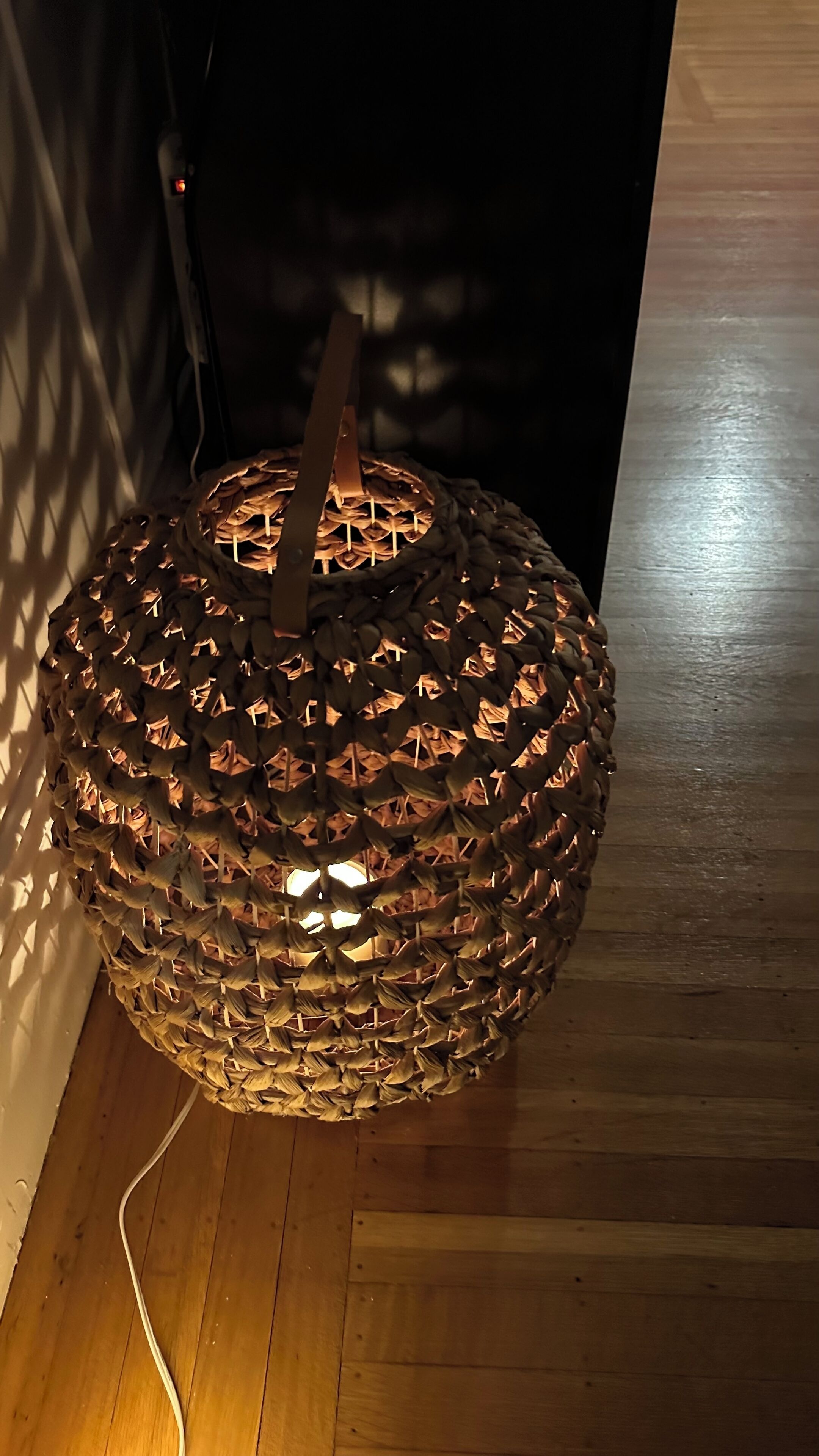 En sfärisk, flätad lampa med ett trähandtag, placerad på ett trägolv intill en vägg.