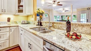 A kitchen with white cabinets, a granite countertop, a double sink, a ceiling fan, and a vase with flowers. - Lakeside Retreat |Private Pool| 8min walk Downtown (Saint Augustine)