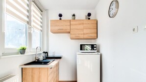 Une cuisine compacte équipée d’un four à micro-ondes, d’un petit réfrigérateur, de meubles en bois, d’une fenêtre avec des stores et d’une horloge murale.
