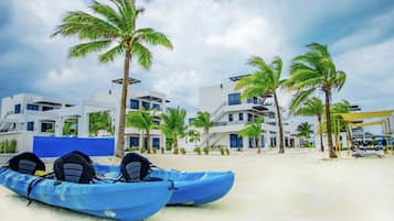 A beach with two blue kayaks, palm trees, and modern buildings with balconies and large windows.