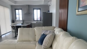 A modern living room with a white leather sectional sofa, a kitchen in the background, and a large window providing natural light. - All Shades of Blu -Best Resort in Caye Caulker (Caye Caulker)
