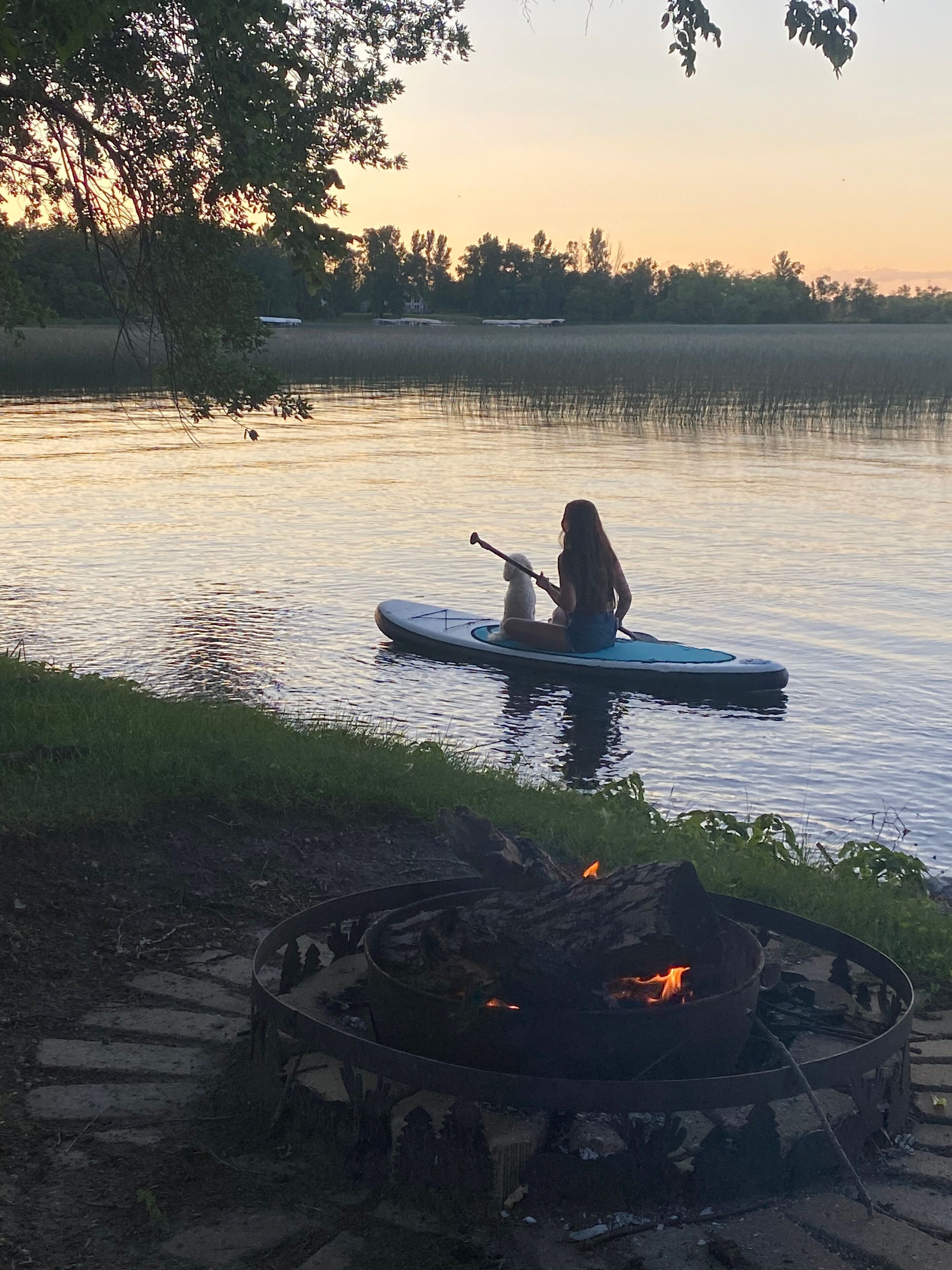 A person paddleboarding on a calm lake with a fire pit in the foreground.