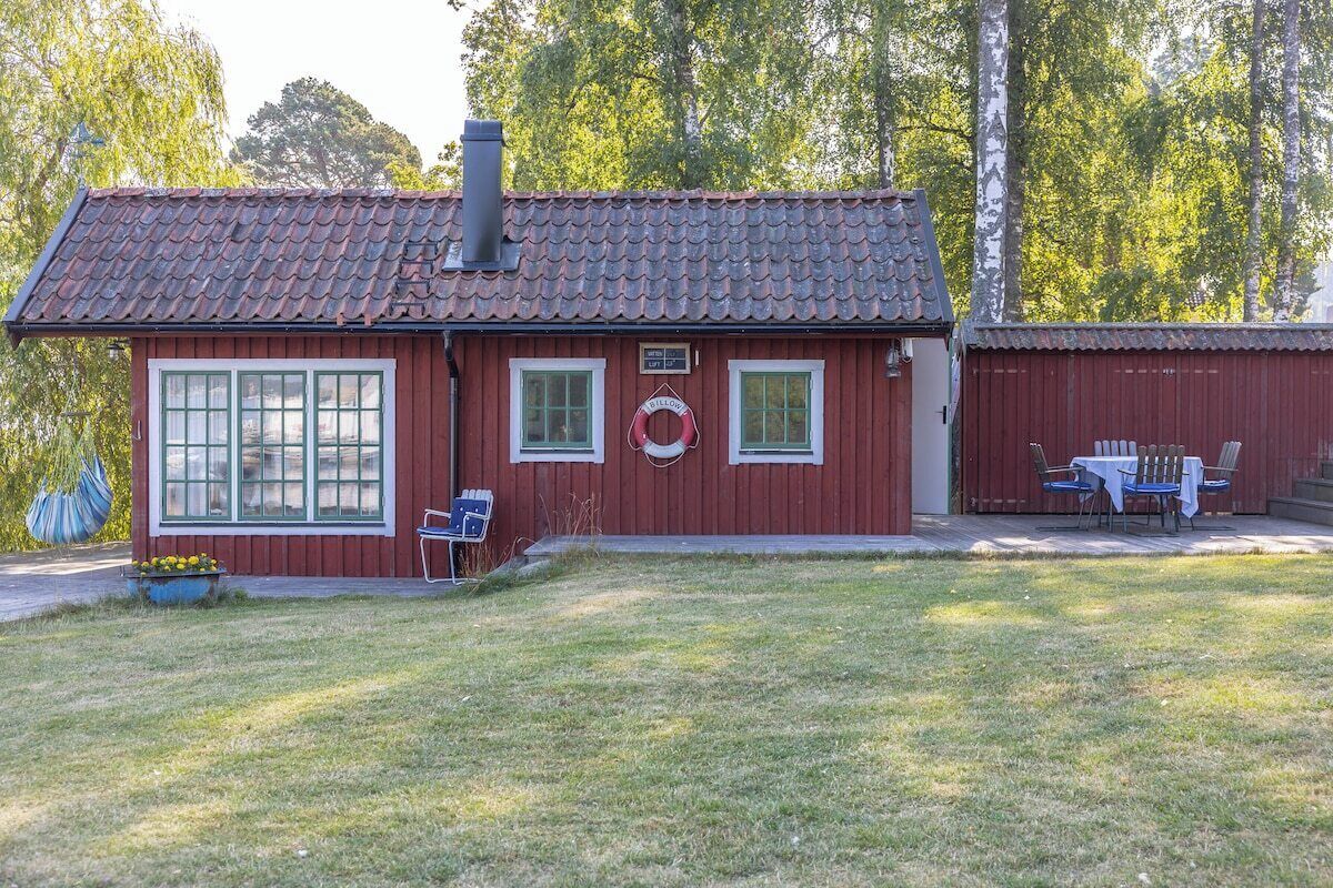 A red wooden cabin with a tiled roof, two windows, and a small porch with a table and chairs.