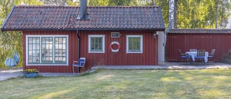 A red wooden cabin with a tiled roof, two windows, and a small porch with a table and chairs.
