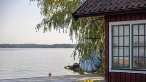 Una cabaña de madera con vista a un lago tranquilo, una hamaca colgada y una maceta con flores amarillas.