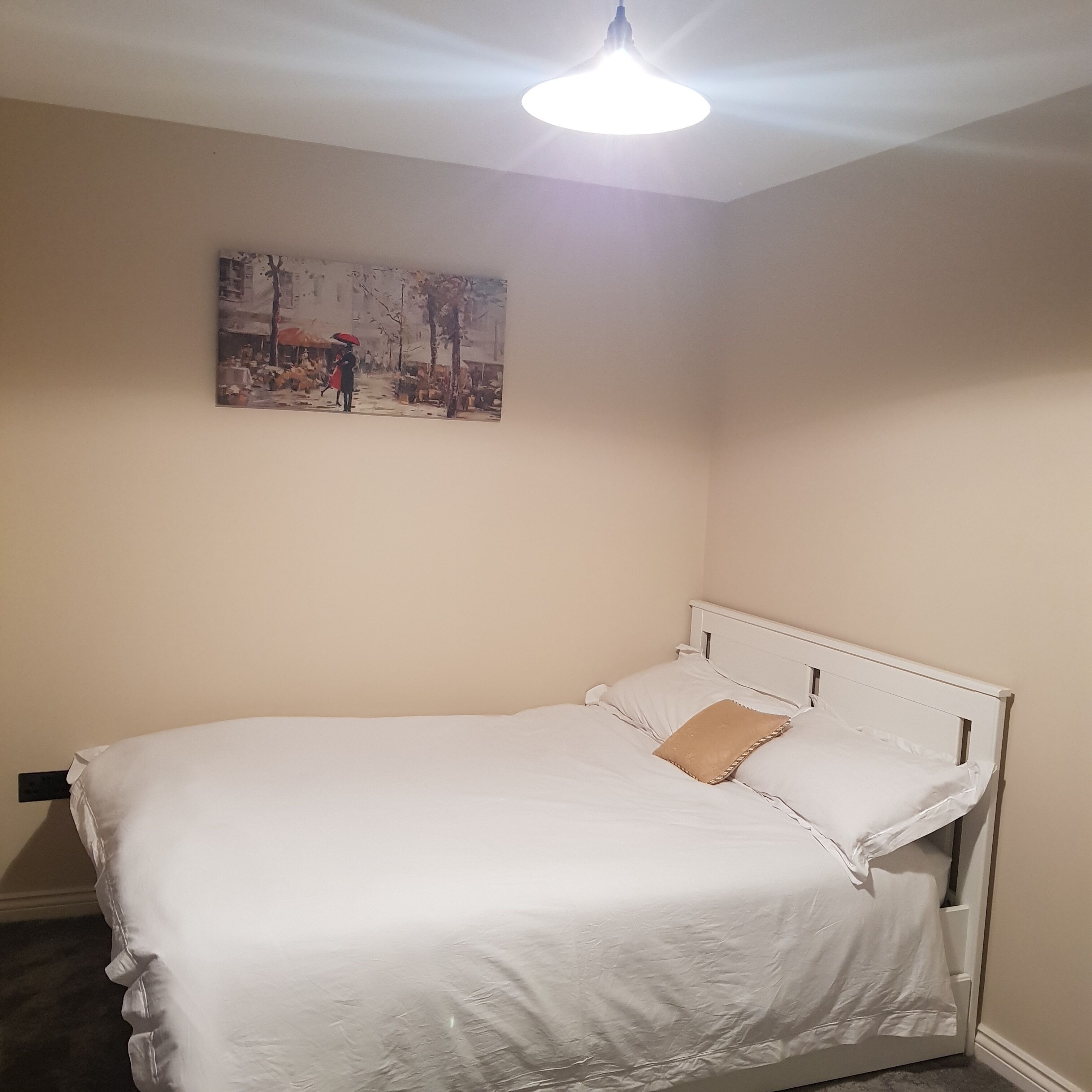 A bedroom with a white bed, a beige wall, and a painting of a rainy street scene.