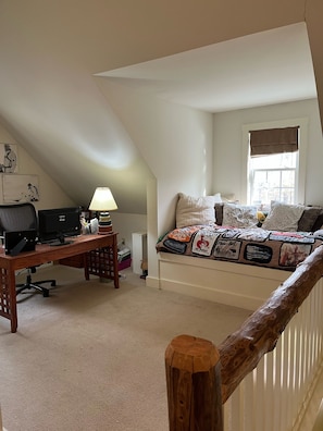 Une chambre avec un plafond en pente, un bureau avec un ordinateur, une lampe et un lit recouvert d’une couverture à motifs.