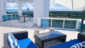 Une terrasse sur le toit, aménagée avec du mobilier d’extérieur, dotée d’une balustrade en verre et offrant une vue sur l’océan et la végétation.