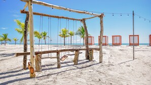 Une balançoire en bois sur une plage de sable, avec des palmiers et un ciel dégagé en arrière-plan.