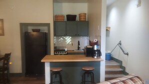 Eine kompakte Küche mit einer zentralen Kochinsel, einem Kühlschrank und einer Kaffeemaschine. Zwei Hocker stehen an der Insel und eine Treppe führt zu einer zweiten Etage.