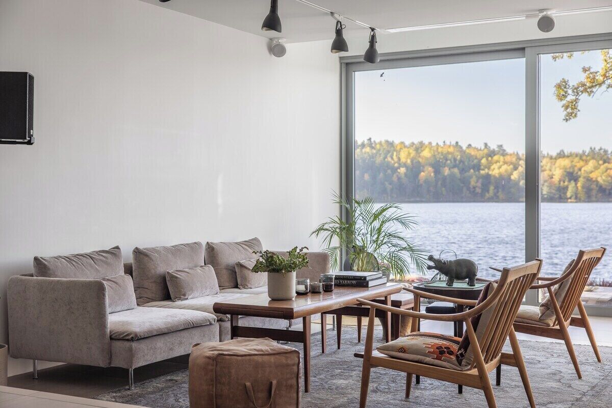 Una sala de estar moderna con un sofá, una mesa de centro y sillas, un ventanal con vistas a un lago y árboles, y un televisor montado en la pared.