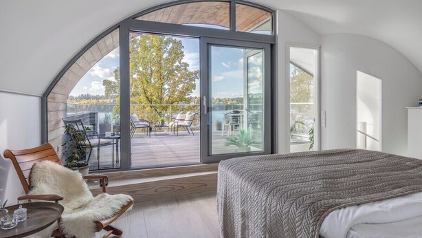 A bedroom with a large arched window, a bed, a rocking chair, and a view of an outdoor deck.