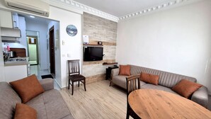 Sala de estar com sofá, mesa de centro de madeira, TV fixada na parede e área de cozinha visível por uma porta aberta.