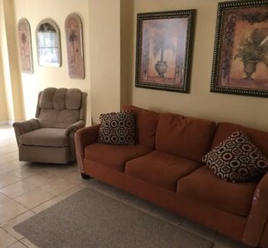 Un salon avec un fauteuil beige, un canapé marron, des coussins à motifs et des œuvres d’art encadrées accrochées aux murs.