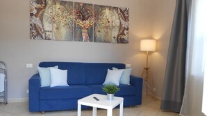 Una sala de estar con un sofá azul, cojines blancos, una mesita auxiliar blanca con una planta y una lámpara de pie.