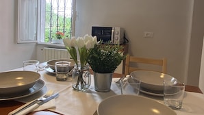 Una mesa de comedor dispuesta con platos, tazones, vasos y cubiertos, junto con un jarrón de flores y una planta en maceta sobre la mesa.