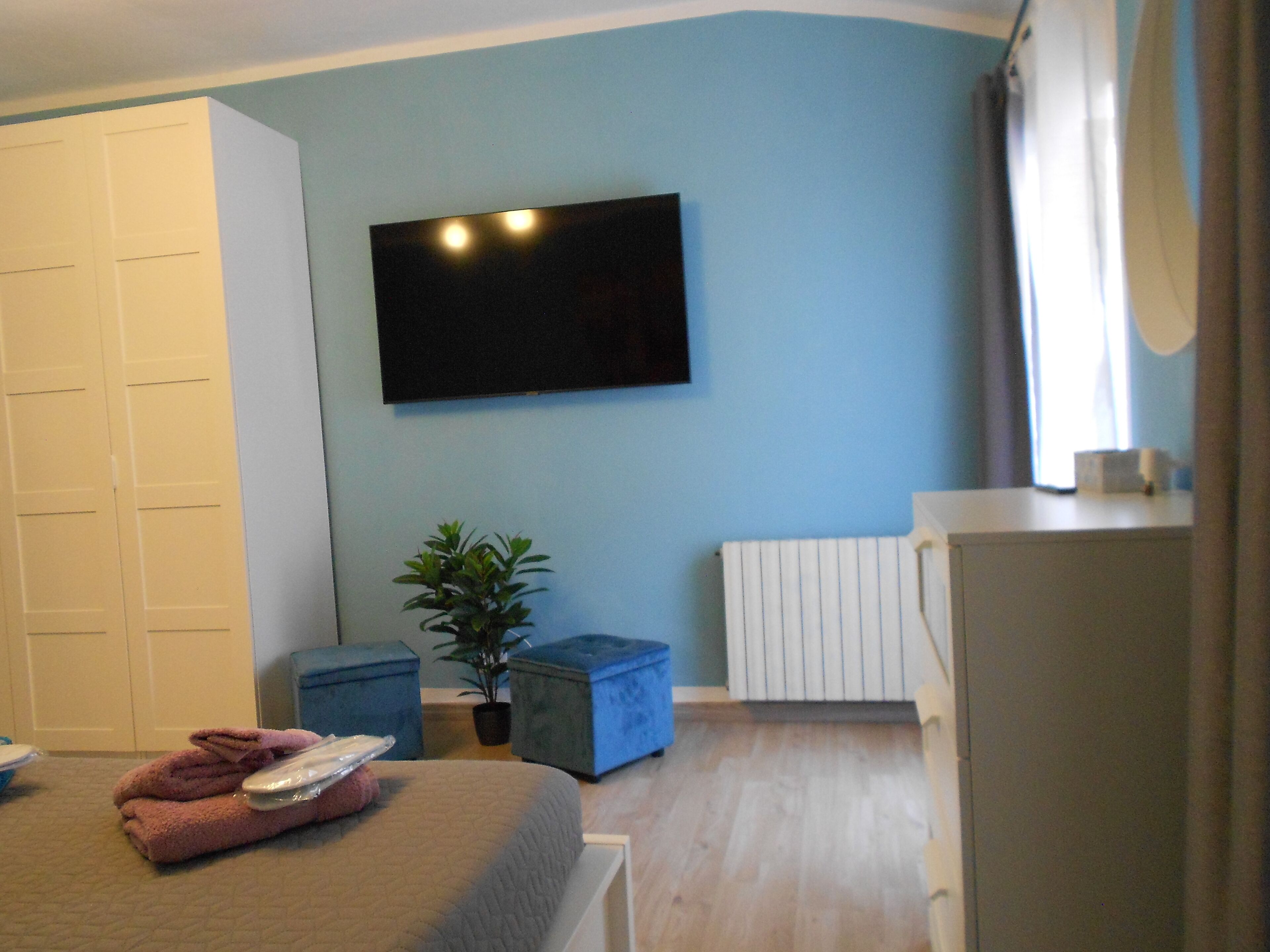 A bedroom with a bed, a TV mounted on the wall, a radiator, and a window with curtains.