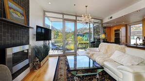 Un moderno salón con chimenea, una mesa de centro de cristal, un sofá blanco y un gran ventanal corredero que da a un balcón con vistas a una casa verde y montañas.