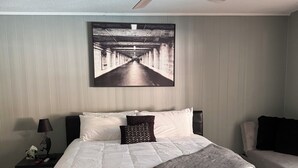 A bedroom with a bed, a nightstand, a lamp, a ceiling fan, and a wall-mounted artwork. - Work Crew Friendly Housing (Tallahassee)