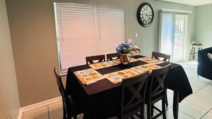 A dining area with a table set for four, a ceiling fan, a clock, and a window with blinds. - Work Crew Friendly Housing (Tallahassee)