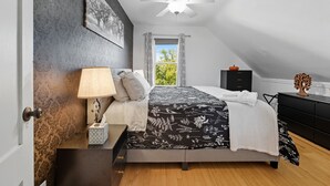 A bedroom with a bed, a nightstand, a lamp, a window with curtains, a ceiling fan, and a dresser.