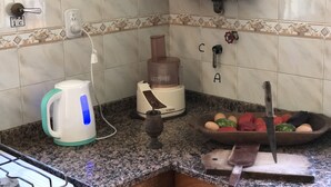 A kitchen with a kettle, a food processor, a cutting board with a knife, and a bowl of fruit.
