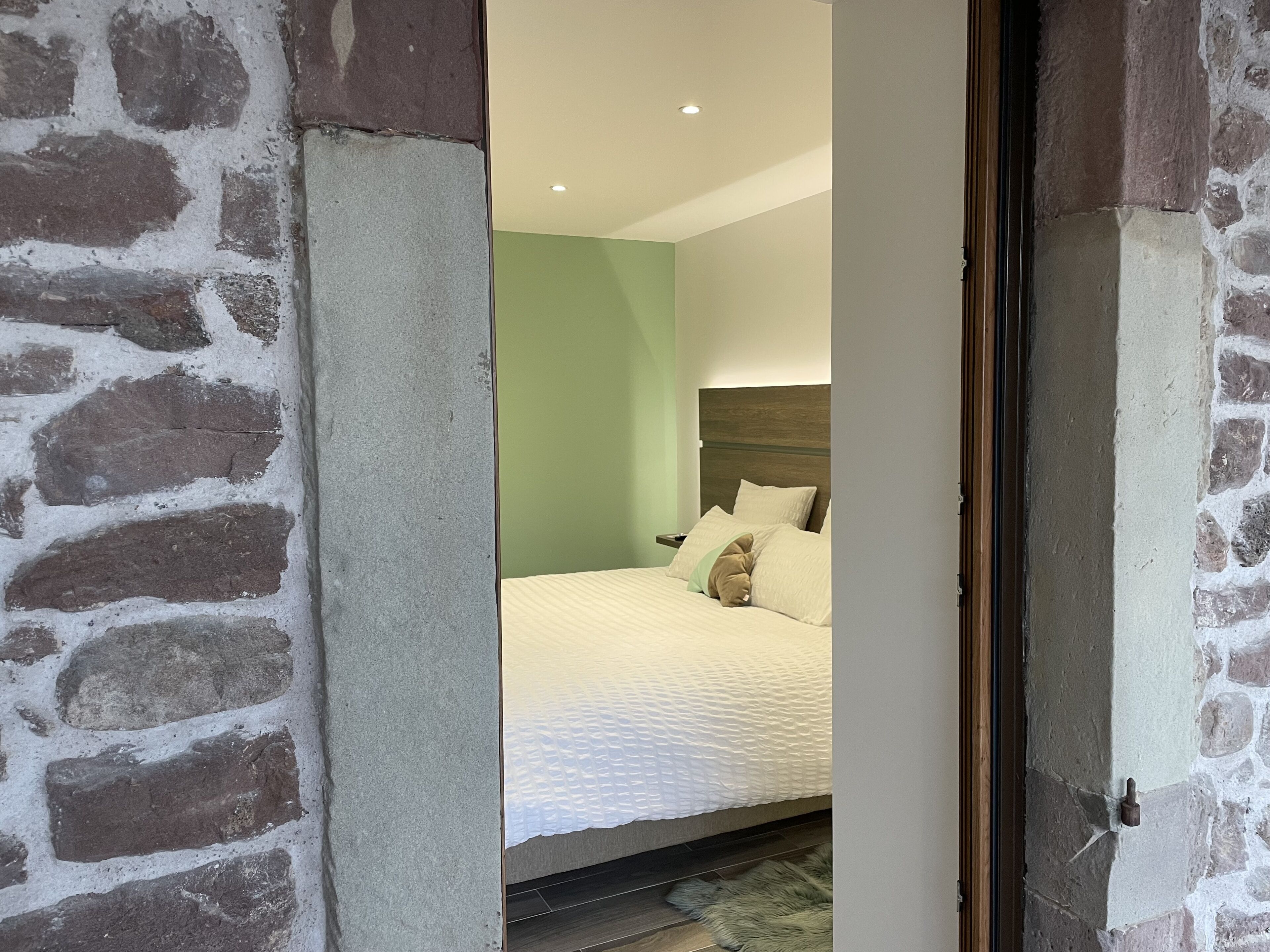 A bedroom with a stone wall, a green wall, a wooden headboard, and a white bedspread.